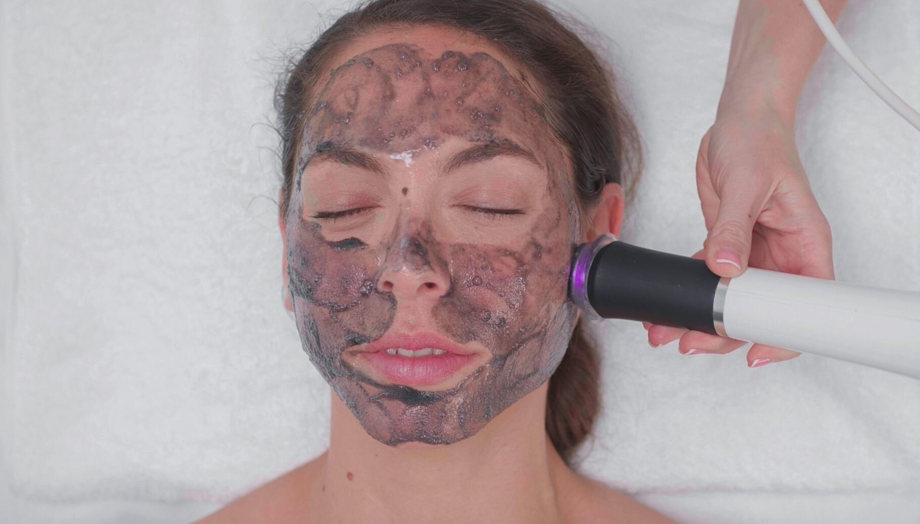 A client relaxing during a facial with a dark treatment mask while a handheld device is moved over her cheek